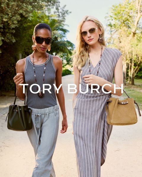 Eine Frau in einem schimmernden Oberteil und einem grünen Rock sitzt neben grünen Geschenkkartons und hält eine geblümte Tory-Burch-Tasche in der Hand; hinter ihr steht ein geschmückter Weihnachtsbaum.