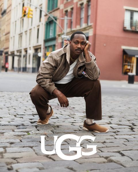 Ein Paar brauner UGG-Wildlederstiefel mit Plüsch-Innenfutter, das auf einer grauen Oberfläche ruht. Das UGG-Logo ist am unteren Rand des Bildes gut sichtbar angebracht.