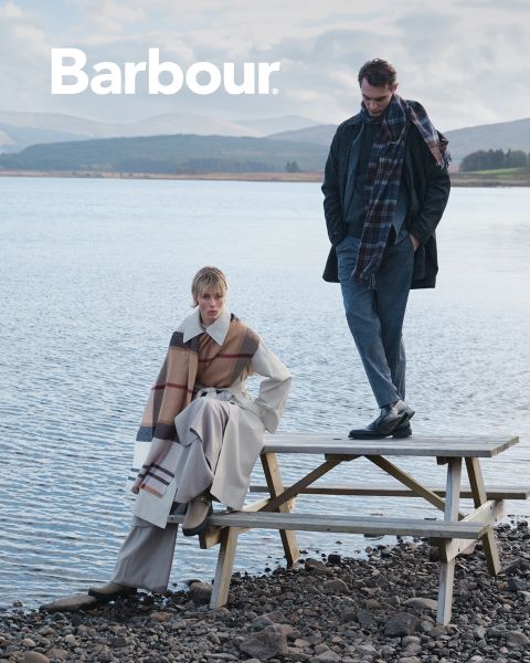 Zwei Personen in stilvoller, mehrlagiger Barbour-Kleidung und karierten Schals posieren an einem See. Einer sitzt auf einem Picknicktisch, während der andere steht, mit Bergen und Wasser im Hintergrund. Das Barbour-Logo erscheint oben links.