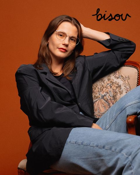 Eine Frau mit Brille, dunklem Blazer und Jeans sitzt auf einem verzierten Stuhl vor einem rostfarbenen Hintergrund, über ihr steht in schwarzer Schrift das Wort „Bisou“.
