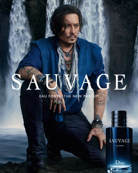 Eine Person mit dunklem Haar und Tattoos kniet vor Wasserfällen. Sie trägt ein blaues Hemd und Schmuck. Das Bild zeigt eine Werbung für „Sauvage“ von Dior. Unten rechts ist eine Flasche des Parfüms zu sehen.