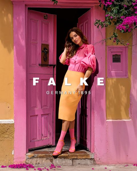 Frau in rosa Bluse und orangefarbenem Rock steht in einem Hauseingang mit rosafarbenen Türen. Sie trägt dazu passende rosafarbene Socken mit orangefarbenen Absätzen. Rosafarbene Blumen zieren die obere Ecke. Weißer Text lautet „FALKE DEUTSCHLAND 1895“.
