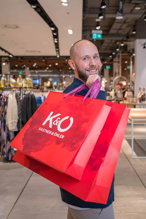 Eine lächelnde Person in einem Geschäft hält zwei große rote Einkaufstüten mit dem Logo von „Kastner & Öhler“. Der Laden ist hell erleuchtet, im Hintergrund stehen Kleiderständer.