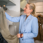 Eine Person in einem hellblauen Blazer lächelt, während sie Kleidung in einem Kleiderschrank betrachtet. Im Fokus steht ein graues Kleidungsstück mit Pailletten. Auf der rechten Seite des Bildes hängt ein brauner Mantel. Die Kulisse scheint ein Kleiderschrank oder ein Ankleidezimmer zu sein.