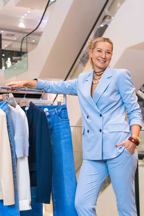 Eine Frau im hellblauen Anzug steht lächelnd neben einem Kleiderständer mit Jeans, Jacken und Oberteilen. Im Hintergrund ist eine Rolltreppe im Innenbereich zu sehen, die eine moderne Einkaufsumgebung schafft.