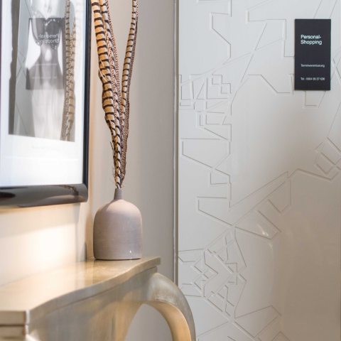 Ein moderner Innenraum mit einer hellen Wand und einem dekorativen Konsolentisch aus Metall. Auf dem Tisch steht eine Vase mit langen, gestreiften Federn. Im Hintergrund ist eine geometrisch gemusterte Tür mit einem „Personal Shopping“-Schild zu sehen.