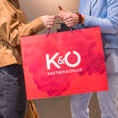 Zwei Personen halten eine große rote Einkaufstasche mit dem Logo „Kastner & Öhler“ und einem rosa Rauchmotiv. Eine Person trägt eine braune Jacke, die andere einen hellblauen Anzug.