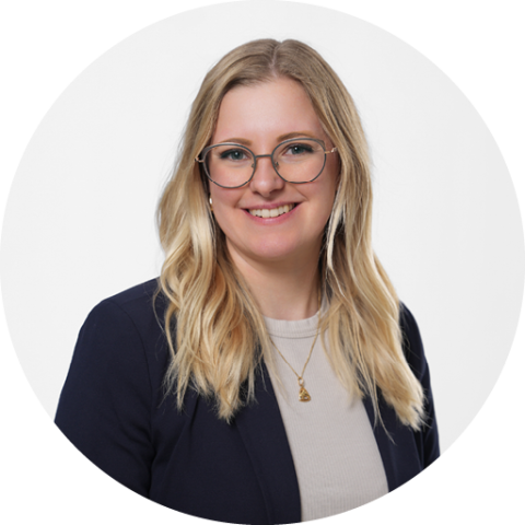 Eine Frau mit langen blonden Haaren, die eine Brille und eine helle Jacke trägt, lächelt vor einem schlichten weißen Hintergrund.