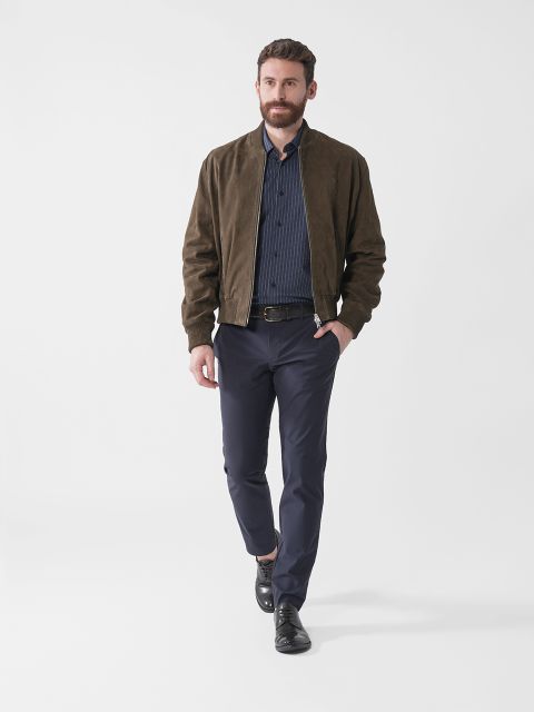 Ein Mann mit Bart kommt näher. Er trägt eine braune Wildlederjacke, ein gestreiftes marineblaues Hemd, eine dunkle Hose und schwarze Schuhe. Der Hintergrund ist schlicht weiß.