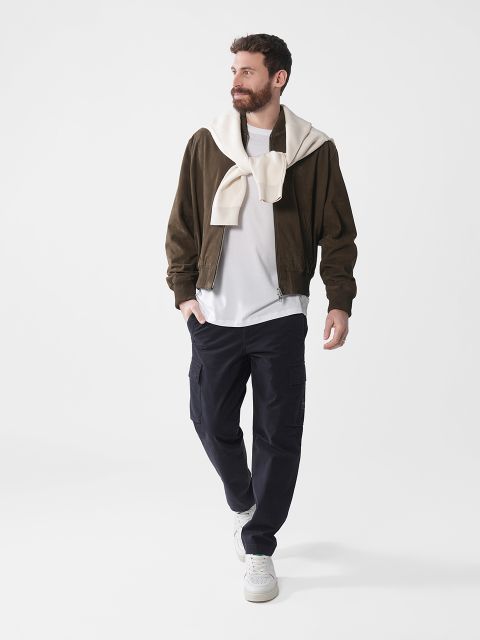 Ein Mann mit Bart geht in dunkler Jacke, weißem Hemd und dunkler Hose. Um seine Schultern ist ein helles Sweatshirt gebunden. Er trägt weiße Turnschuhe und ist von neutraler Beleuchtung und einem schlichten Hintergrund umgeben.