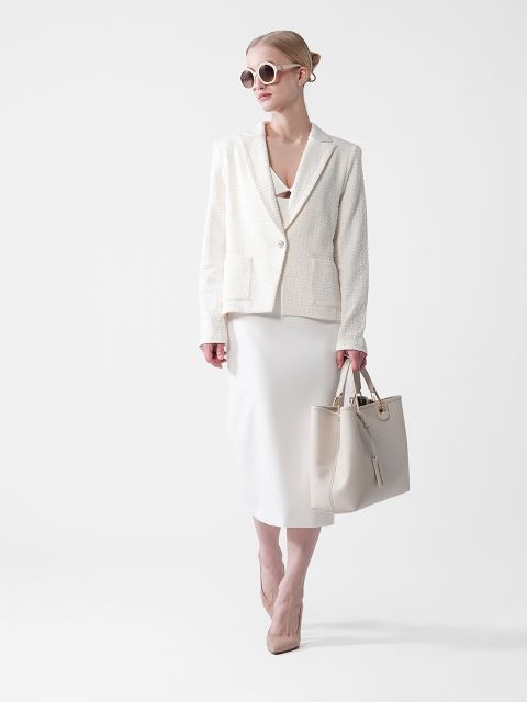 Eine Frau mit stylischer Sonnenbrille trägt einen cremefarbenen Blazer und Rock und hält eine große beige Handtasche in der Hand. Sie steht selbstbewusst vor einem schlichten weißen Hintergrund und verbindet Eleganz und moderne Mode.