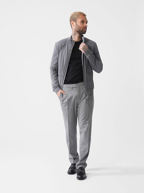 Ein Mann mit kurzen Haaren und Tattoos trägt eine graue Jacke, ein schwarzes Hemd und eine graue Hose und steht vor einem schlichten weißen Hintergrund. Er hat eine Hand in der Tasche und blickt zur Seite.