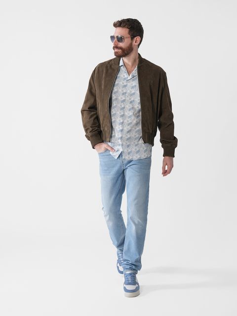Ein Mann mit Bart, Sonnenbrille, brauner Jacke, gemustertem Hemd, hellblauen Jeans und Turnschuhen geht selbstbewusst vor einem schlichten weißen Hintergrund.