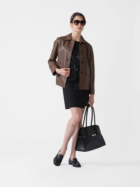 Eine Person, die eine braune Lederjacke über einem schwarzen Kleid und eine Sonnenbrille trägt, steht selbstbewusst vor einem schlichten Hintergrund. Sie trägt eine schwarze Handtasche und schwarze Slipper.