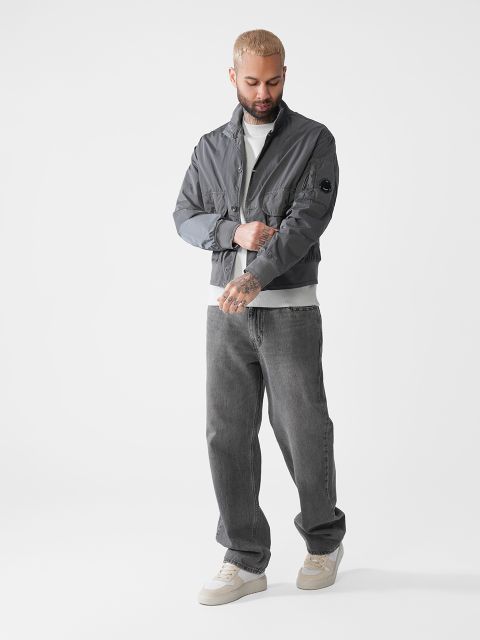 Ein Mann mit kurzen Haaren und Tattoos steht vor einem schlichten weißen Hintergrund. Er trägt eine graue Jacke, einen weißen Rollkragenpullover, dunkelgraue Jeans und weiße Turnschuhe. Er blickt nach unten und berührt sein Handgelenk.