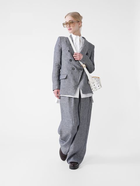 Eine Person in einem eleganten grauen Anzug mit weiten Hosenbeinen und einem weißen Hemd geht selbstbewusst. Sie trägt eine große Sonnenbrille, hält eine kleine weiße Handtasche in der Hand und hat die Haare zurückgekämmt. Der Hintergrund ist schlicht weiß.