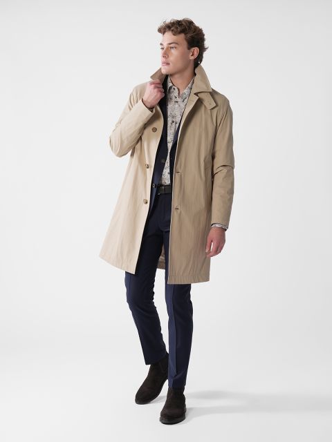 Eine Person trägt einen hellbraunen Trenchcoat über einem gemusterten Hemd und einer dunklen Hose vor einem schlichten weißen Hintergrund. Sie blickt zur Seite, berührt mit einer Hand den Mantelkragen und trägt dunkle Schuhe.
