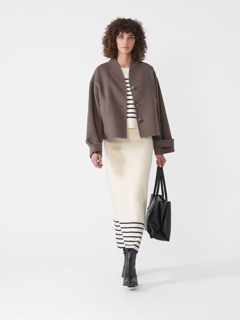 Eine Person mit lockigem Haar läuft selbstbewusst in einer braunen Jacke über einem gestreiften creme-schwarzen Kleid. Sie trägt eine große schwarze Einkaufstasche und schwarze Stiefel vor einem schlichten weißen Hintergrund.