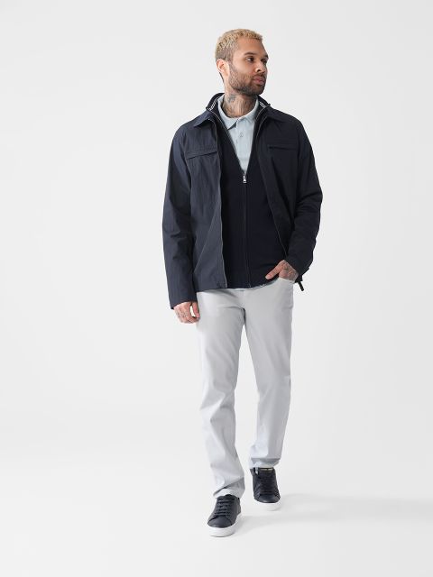 Ein Mann mit hellem Haar und Tattoos steht vor einem schlichten weißen Hintergrund. Er trägt eine schwarze Jacke, ein hellblaues Hemd, eine hellgraue Hose und schwarze Turnschuhe. Er blickt entspannt zur Seite.