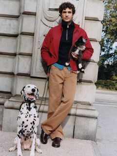Ein Mann mit lockigem Haar lehnt an einer Steinmauer und hält eine Katze im Arm. Er trägt eine rote Jacke, einen schwarzen Pullover, ein blaues Hemd und beige Hosen. Neben ihm sitzt ein Dalmatiner an der Leine. Im Hintergrund sind Bäume und ein Fußgängerüberweg zu sehen.