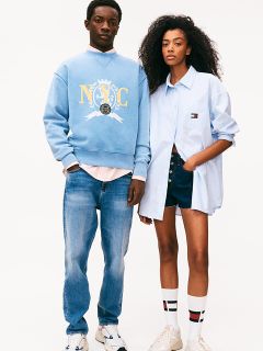 Ein Mann und eine Frau stehen nebeneinander vor einem weißen Hintergrund. Der Mann trägt ein blaues NYC-Sweatshirt, helle Jeans und Turnschuhe. Die Frau trägt ein hellblaues Hemd, Jeansshorts und Kniestrümpfe. Beide haben eine lässige, entspannte Haltung.