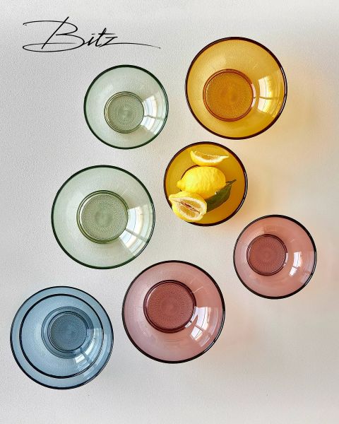Sieben bunte Glasschalen, kreisförmig angeordnet auf einer weißen Fläche. Eine gelbe Schale enthält drei Zitronen mit grünen Blättern. Das "Bitz"-Logo ist in der oberen linken Ecke zu sehen.