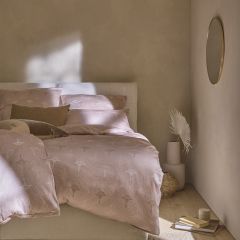 Ein gemütliches Schlafzimmer mit einem beigefarbenen Bett mit gemusterter Bettwäsche. An der Wand hängt ein runder Spiegel. Sanftes Licht beleuchtet einen Teil des Bettes. Ein kleiner Stapel Bücher und Dekorationsgegenstände liegen in der Nähe auf dem Boden.