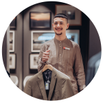Eine Person in einem braunen Hemd hält einen Kleiderbügel mit einem beigen Blazer. Sie lächeln und stehen in einem Raum mit gerahmten Bildern an den Wänden.