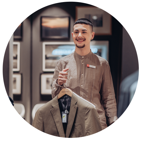 Eine Person in einem braunen Hemd hält einen Kleiderbügel mit einem beigen Blazer. Sie lächeln und stehen in einem Raum mit gerahmten Bildern an den Wänden.