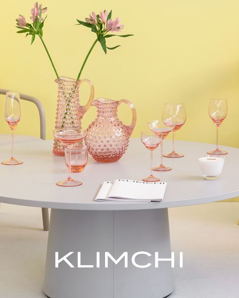 Ein stilvoll gedeckter Tisch besteht aus rosafarbenem Glasgeschirr, darunter Krüge, Gläser und Kelche. Rosafarbene Blumen in einer Vase verleihen einen Hauch von Eleganz. Der Hintergrund ist in sanftem Gelb gehalten, und am unteren Bildrand ist das Wort „KLIMCHI“ zu sehen.