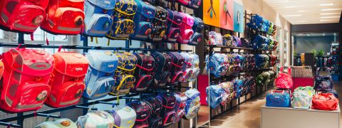 Eine hell erleuchtete Ladenauslage präsentiert eine große Auswahl an bunten Rucksäcken in Regalen. Die Taschen sind in verschiedenen Farben erhältlich, darunter Rot, Blau, Pink und mehr, und haben unterschiedliche Designs. Der Raum verfügt über weiße Deckenbeleuchtung und ein modernes Interieur.