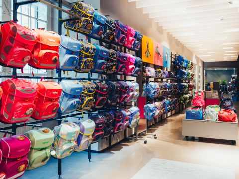 Eine hell erleuchtete Ladenauslage präsentiert eine große Auswahl an bunten Rucksäcken in Regalen. Die Taschen sind in verschiedenen Farben erhältlich, darunter Rot, Blau, Pink und mehr, und haben unterschiedliche Designs. Der Raum verfügt über weiße Deckenbeleuchtung und ein modernes Interieur.