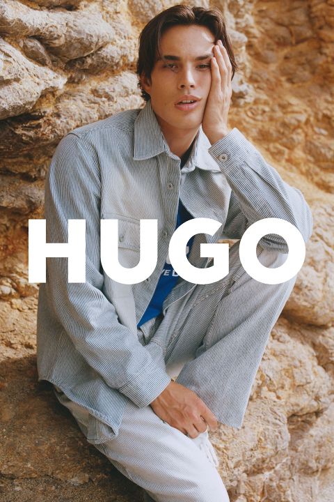 Eine Person sitzt vor einer felsigen Kulisse. Sie trägt ein hell gestreiftes Outfit und ein blaues Hemd. Sie legt eine Hand auf ihr Gesicht. Das Wort „HUGO“ ist deutlich sichtbar auf dem Bild zu sehen.