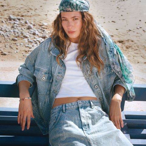Eine Person sitzt auf einer Bank am Strand. Sie trägt eine Jeansjacke und Jeans mit einem weißen Oberteil. Sie hat lange Haare und trägt ein grün gemustertes Bandana. Im Hintergrund sind Kieselsteine und Sand zu sehen.