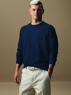 Eine Person mit kurzen, hellen Haaren steht selbstbewusst in einem Studio. Sie trägt einen dunkelblauen Rippenpullover und eine helle Hose mit passendem Gürtel. Die Hände stecken in den Taschen, der Gesichtsausdruck ist neutral.
