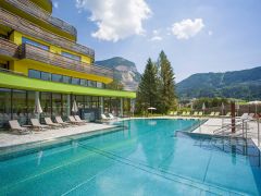 Modernes Hotel mit geschwungener Architektur und türkisfarbenem Pool im Vordergrund, umgeben von Liegestühlen. Berglandschaft und blauer Himmel mit Wolken im Hintergrund.