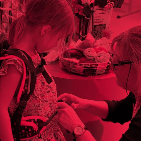Eine Frau hilft einem Kind in einem Geschäft, die Riemen eines Rucksacks zu richten. Die Szene ist rot gefärbt. Sie stehen neben einem Regal mit Aufklebern und Spielzeug. Das Kind beobachtet aufmerksam.