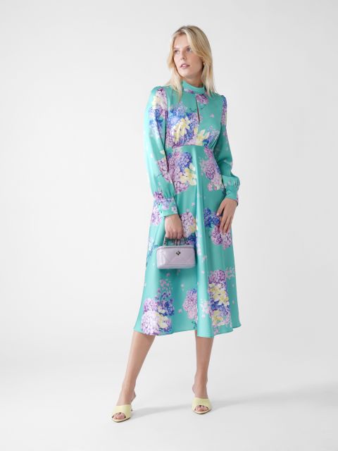 Eine Person in einem pastellblauen Blumenkleid posiert vor einem schlichten Hintergrund. Sie hält eine kleine lavendelfarbene Handtasche und trägt hellgelbe Sandalen mit Absatz. Das Kleid hat lange Ärmel und einen hohen Kragen.