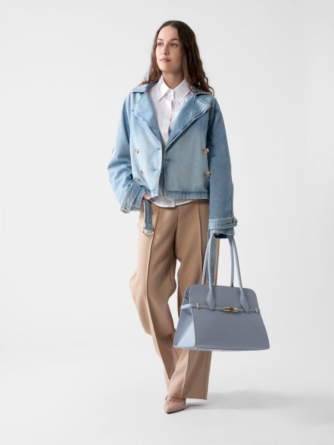 Eine Person mit langen Haaren steht vor einem schlichten Hintergrund. Sie trägt eine blaue Jeansjacke über einem weißen Hemd und einer beigen Hose. Sie hält eine große hellblaue Handtasche in der Hand und blickt in die Ferne.