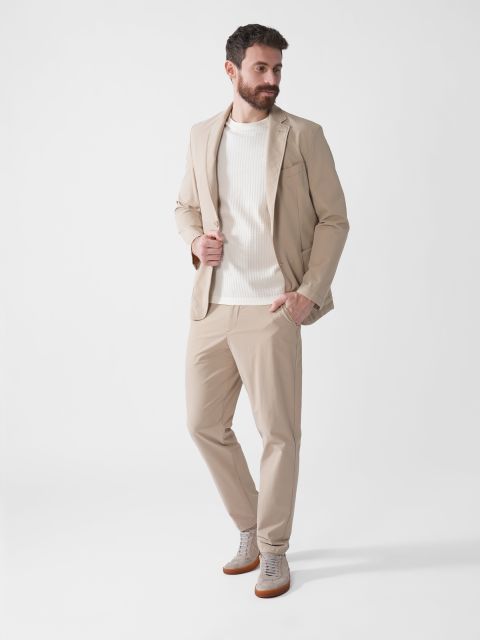 Ein Mann mit Bart steht da und trägt eine beige Jacke, eine Hose und einen weißen Strickpullover. Er blickt zur Seite und hat eine Hand in der Jackentasche. Dazu trägt er helle Turnschuhe. Der Hintergrund ist schlicht weiß.