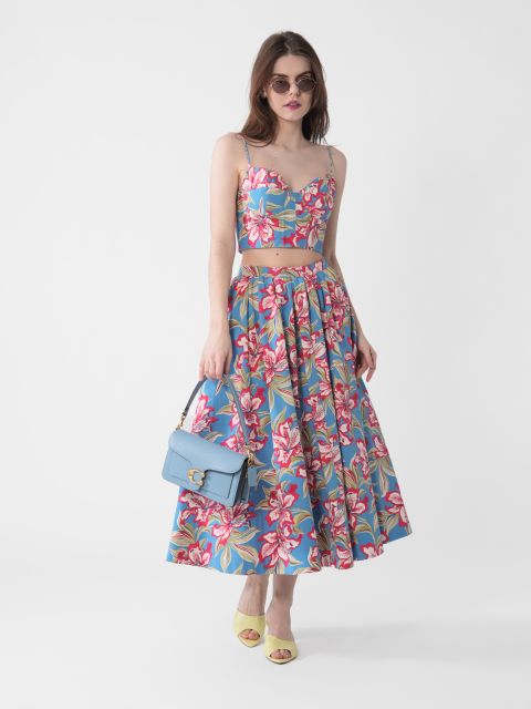 Frau mit Sonnenbrille, geblümtem Crop-Top und passendem Rock mit rosa-weißen Blumen auf blauem Grund. Sie hält eine hellblaue Handtasche in der Hand und trägt gelbe offene Schuhe. Sie steht vor einem schlichten weißen Hintergrund.
