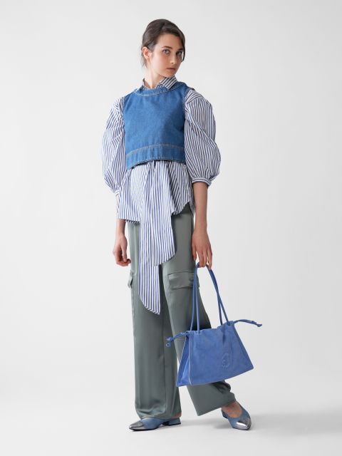Eine Person steht in einem Studio. Sie trägt eine blaue Jeansweste über einer gestreiften Bluse mit Puffärmeln, graue Cargohosen und blaue Schuhe. Sie hält eine blaue Wildlederhandtasche in der Hand. Der Hintergrund ist schlicht weiß.