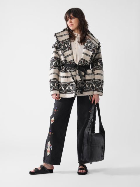 Eine Person posiert in einer gemusterten Jacke mit Fransendetails, einem weißen Hemd und einer schwarzen Hose mit bunten Stickereien. Sie trägt eine schwarze Tasche und schwarze Sandalen und steht vor einem schlichten weißen Hintergrund.