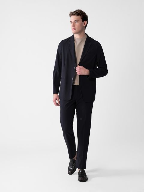 Eine Person im Smart-Casual-Outfit posiert vor einem schlichten weißen Hintergrund. Sie trägt einen dunklen Blazer, eine dazu passende Hose, ein helles Hemd und schwarze Schuhe, blickt zur Seite und hat eine Hand in der Hosentasche.