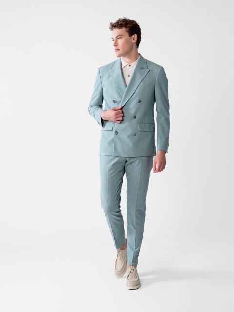 Eine Person in einem hellblauen Zweireiher und einem weißen Hemd mit Kragen läuft selbstbewusst vor einem schlichten weißen Hintergrund. Sie trägt beige Schuhe und hat kurze, lockige Haare. Ihr Blick ist nach rechts gerichtet.