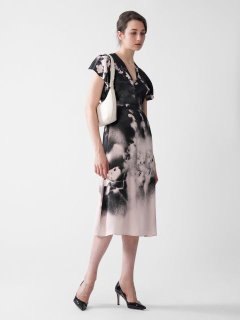 Eine Frau in einem rosa-schwarz gemusterten Kleid steht vor einem hellen Hintergrund. Sie hält eine weiße Handtasche in der Hand und trägt schwarze High Heels. Ihr Haar ist ordentlich frisiert, und sie wirkt selbstbewusst und elegant.