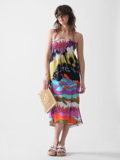Frau mit Sonnenbrille und buntem, gemustertem Sommerkleid mit Neckholder-Trägern, hält eine gewebte Clutch und steht in Sandalen vor einem schlichten weißen Hintergrund.