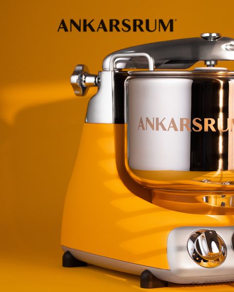 Nahaufnahme einer gelben Ankarsrum-Küchenmaschine mit einer glänzenden Rührschüssel aus Edelstahl vor einem passenden gelben Hintergrund. Der Markenname Ankarsrum erscheint über der Maschine.