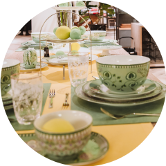 Festlich gedeckter Tisch mit grün gemusterten Tellern und Tassen auf grünen Tischsets und einer gelben Tischdecke. Ein Etagerenständer mit grünen und gelben Deko-Eiern dient als Mittelpunkt. Verschiedene Besteck- und Glaswaren runden das Ambiente ab.