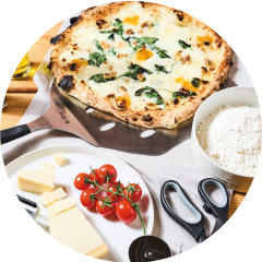 Eine frisch gebackene Pizza mit Spinat und Nüssen liegt auf einem Pizzaschieber. Um sie herum liegen Zutaten wie Kirschtomaten, Käse und eine Schüssel Mehl. Schere und ein Flaschenöffner liegen auf dem Tisch.